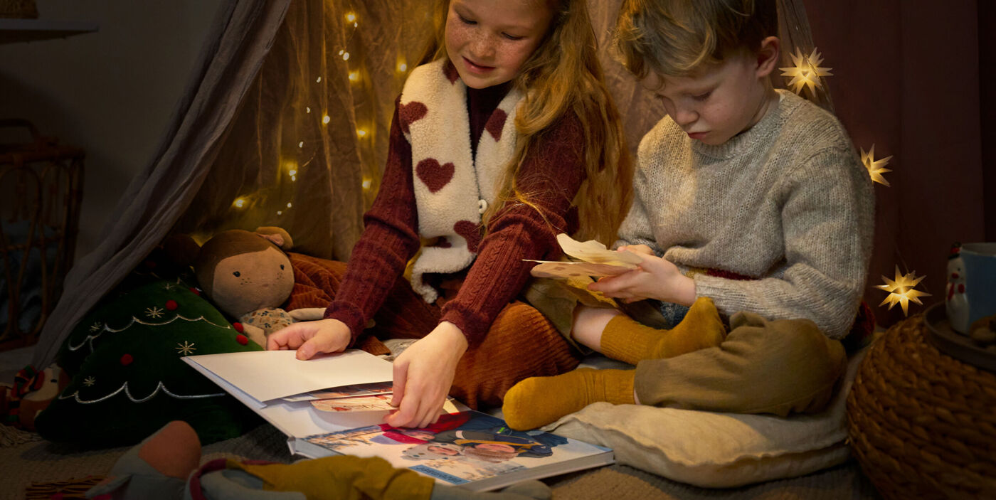 Zwei Kinder betrachten gemeinsam Erinnerungen aus einer Erinnerungstasche in einem CEWE Fotobuch in gemütlicher Weihnachtsstimmung.