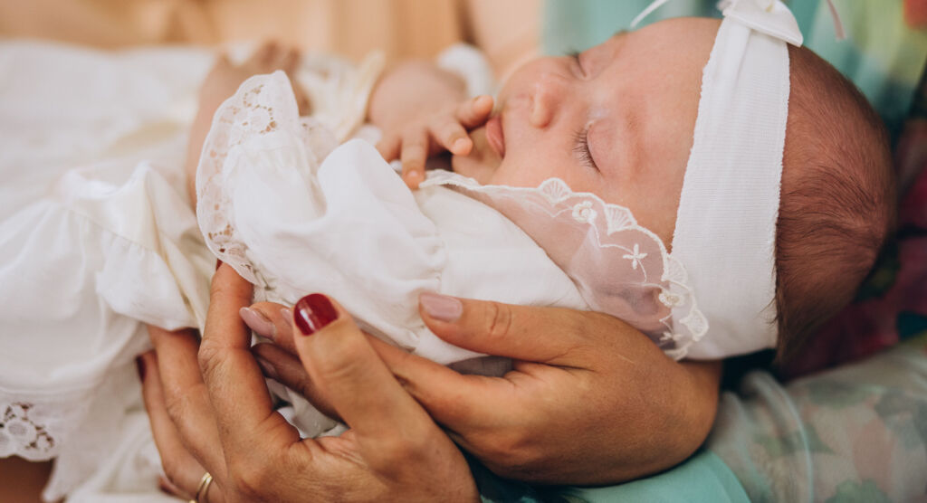 Neugeborenes Baby in weißer Kleidung und mit Stirnband wird von einer erwachsenen Person gehalten.