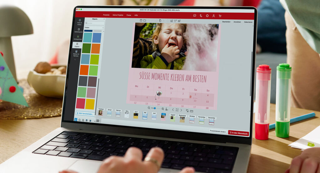 Eine Frau sitzt vor einem Laptop und gestaltet einen Fotokalender. Im Hintergrund ist ein spielendes Kind zu sehen.