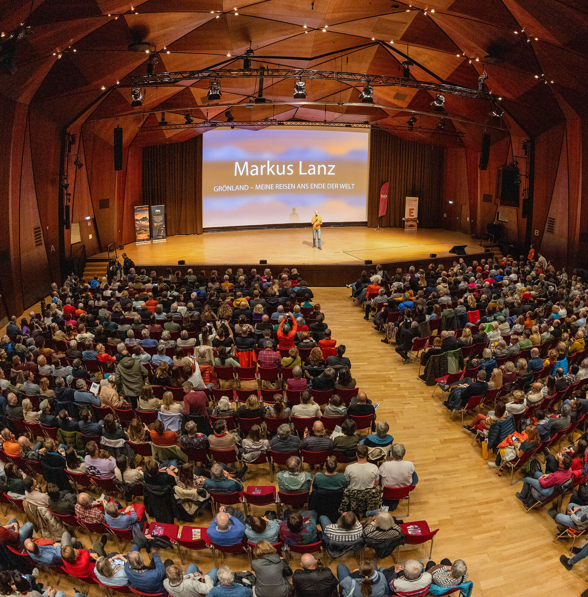 In einem großen Saal sitzen viele Menschen in Stuhlreihen vor einer Leinwand. Auf der Bühne steht ein Mann und hält einen Vortrag.